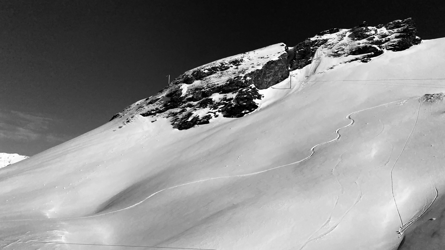 Black-and-white view of curved ski tracks across a snow-covered mountainside beneath a rocky ridge