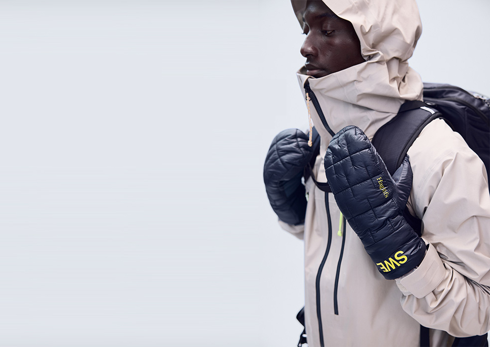 A model holding backpack straps while wearing navy Haglöfs Team SWE mittens and a light shell jacket.