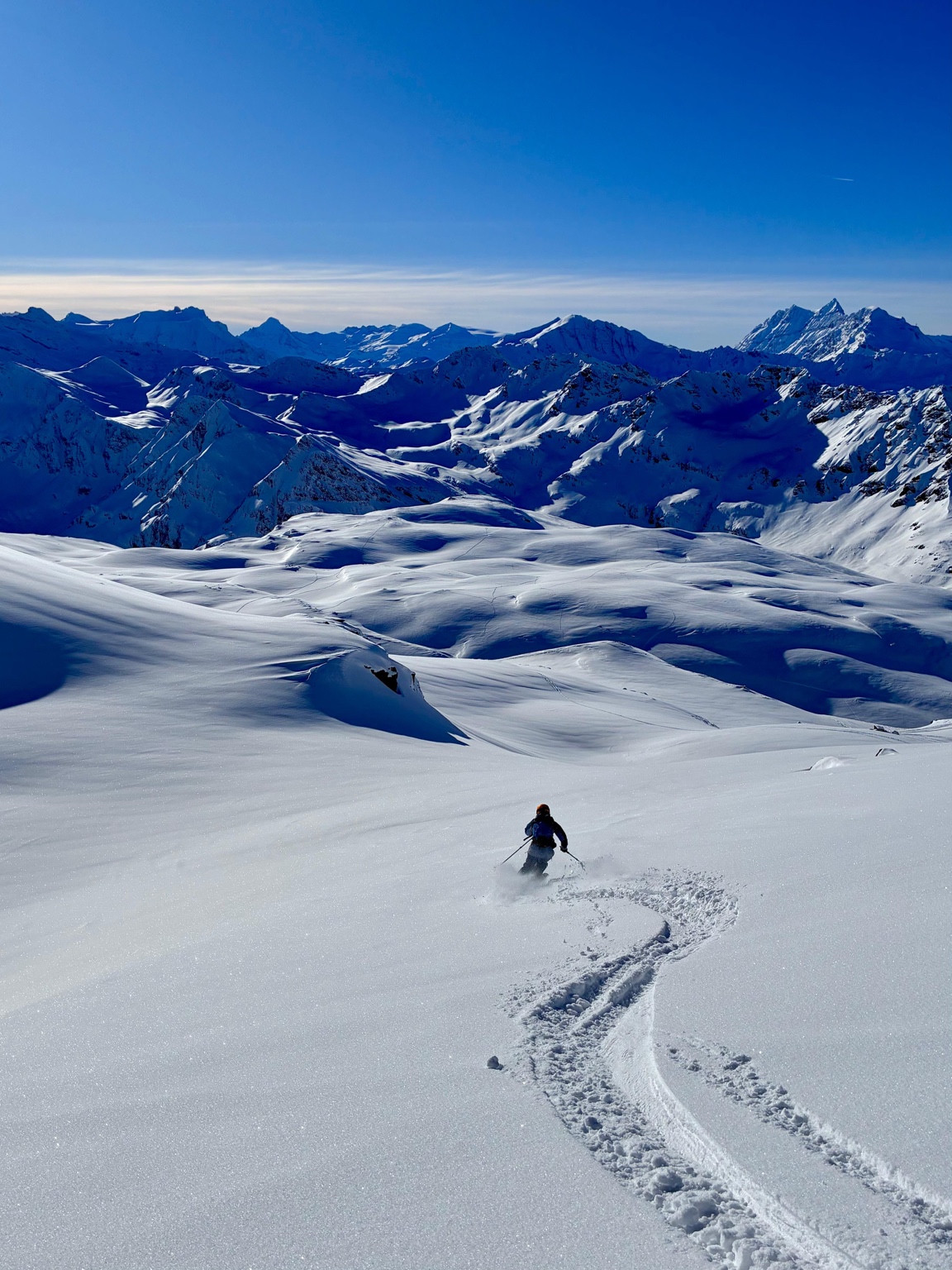 A skier carving through powder on a broad snow-covered slope, with curved tracks in the foreground and mountain ridges beneath a blue sky